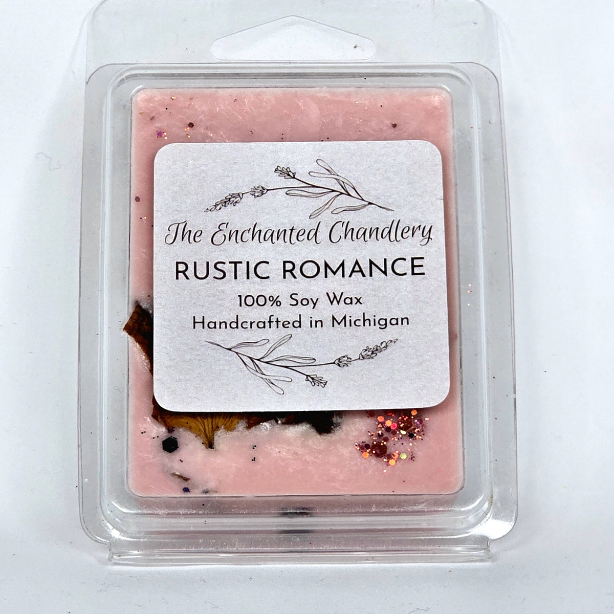 A single square wax melt in a clear plastic clamshell package on a white background. The wax is light pink with small scattered botanical and glitter inclusions visible through the clear container.